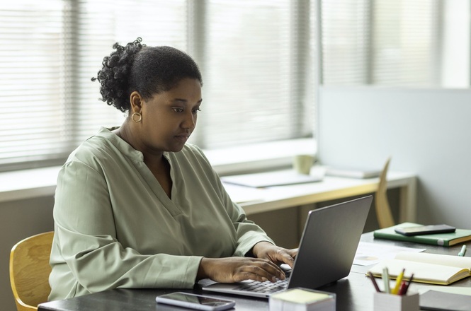 A imagem mostra uma mulher de pele escura sentada em uma mesa de escritório, usando um laptop. Ela está concentrada olhando para a tela enquanto digita. A mulher tem cabelo encaracolado preso em um coque e está vestindo uma blusa verde clara. No fundo, há uma janela com persianas e uma mesa com alguns objetos. Na mesa em que ela está trabalhando, há também um smartphone e um porta-lápis.