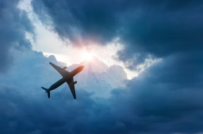 A imagem mostra um avião voando no céu, visto de baixo. O avião está em silhueta, contrastando com o céu ao fundo. O céu está parcialmente nublado, com nuvens escuras, mas há uma abertura entre as nuvens por onde está a luz do sol.