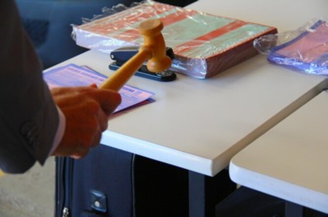 A imagem mostra uma mão segurando um martelo de leilão, prestes a bater em uma superfície branca, que parece ser uma mesa. Ao fundo, sobre a mesa, há alguns objetos embalados em plástico transparente, incluindo o que parece ser um livro ou caderno grande e um estojo ou pasta de cor azul.