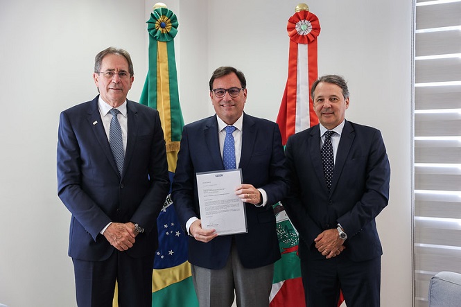 A imagem mostra três homens de terno e gravata posando para uma foto em um ambiente interno. Eles estão em pé, lado a lado. O Desembargador Francisco Oliveira Neto está no centro segurando um documento em suas mãos. Ao lado dele estão os representantes da Fiesc  Há duas bandeiras ao fundo: a do Brasil à esquerda e a de Santa Catarina à direita. O ambiente parece ser um escritório ou sala de reuniões, com uma janela com persianas à direita.