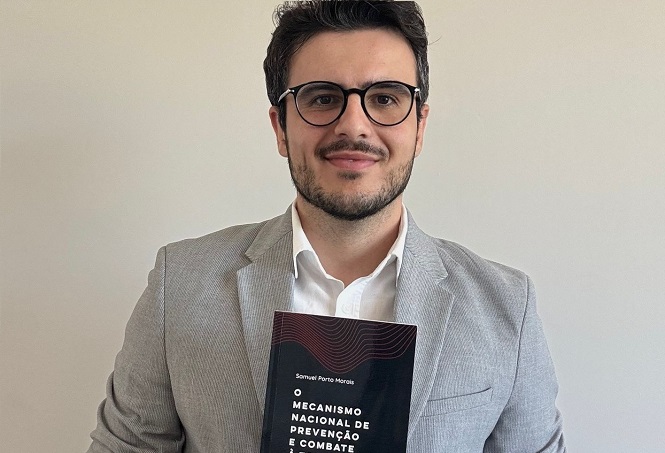 A imagem mostra um homem de pele clara, cabelo castanho escuro e barba curta. Ele está usando óculos de armação preta e veste um terno cinza claro com uma camisa branca por baixo. O homem está segurando um livro na frente do peito. A capa do livro é predominantemente preta com detalhes em vermelho. O título do livro é "O MECANISMO NACIONAL DE PREVENÇÃO E COMBATE". O nome do autor, "Samuel Porto Morais", está escrito acima do título. O fundo da imagem é uma parede branca lisa.