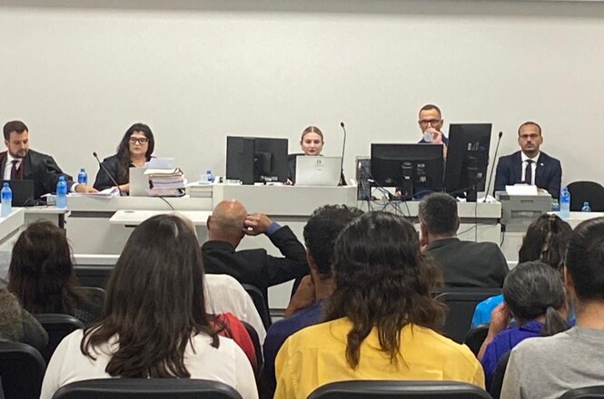 A imagem mostra uma sala de audiência ou tribunal. Na parte da frente, há uma mesa comprida com cinco pessoas sentadas atrás dela, todas vestidas formalmente. Na frente da mesa, há várias pessoas sentadas de costas para a câmera, assistindo à audiência. A sala parece ser um ambiente formal e profissional.