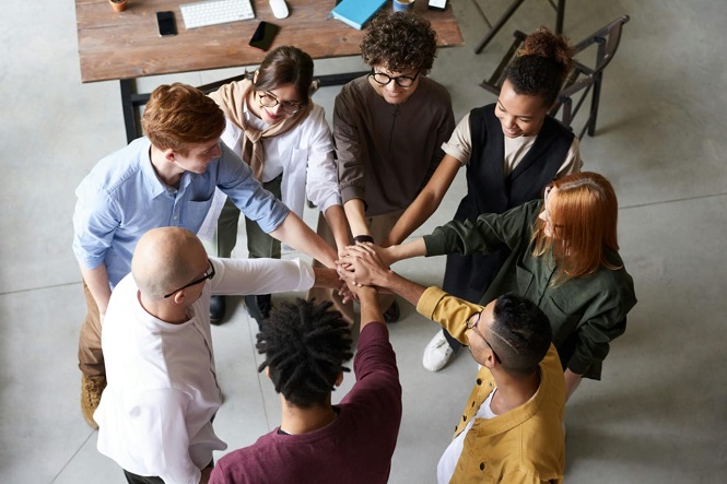 A imagem mostra um grupo de oito pessoas em pé formando um círculo. Elas estão com as mãos estendidas para o centro, sobrepondo-as umas sobre as outras em um gesto de união ou trabalho em equipe. As pessoas são diversas em termos de gênero e etnia. Ao fundo, há uma mesa de madeira com alguns objetos, como um computador, cadernos e canetas.