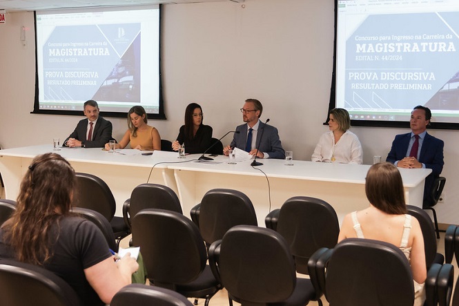 A imagem mostra uma mesa de conferência com seis pessoas sentadas, todas vestidas formalmente. Elas estão em uma sala de conferências, com cadeiras vazias na frente e algumas pessoas sentadas assistindo. Atrás dos palestrantes, há duas telas de projeção exibindo a mesma imagem, que contém o texto: "Concurso para ingresso na Carreira da Magistratura Edital n. 44/2024 Prova Discursiva Resultado Preliminar". A sala parece ser um ambiente formal, possivelmente em um evento relacionado a concursos públicos para a magistratura.