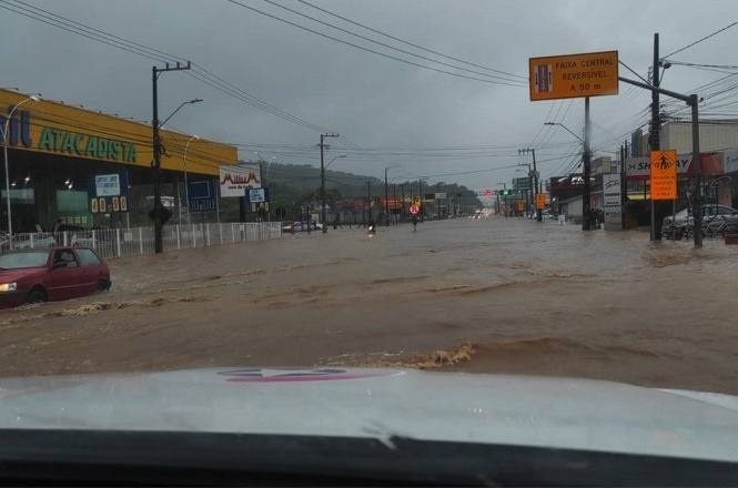 A imagem mostra uma rua completamente alagada, com água cobrindo quase toda a via. Há um carro vermelho parcialmente submerso à esquerda da imagem. À direita, há placas de trânsito, uma delas indicando "Faixa Central Reversível a 50 m". No lado esquerdo, há um estabelecimento comercial com uma fachada amarela e azul com o nome "Atacadista". O céu está nublado, indicando que o tempo está chuvoso. A cena parece ser de uma área urbana, com postes de iluminação e fiação elétrica visíveis.