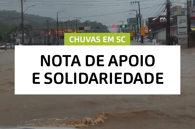A imagem mostra uma rua alagada devido a fortes chuvas. No topo da imagem, há um retângulo verde com o texto "CHUVAS EM SC". Abaixo, em um fundo branco, está escrito "NOTA DE APOIO E SOLIDARIEDADE". Ao fundo, é possível ver postes de eletricidade e fachadas de lojas parcialmente cobertas pela água.