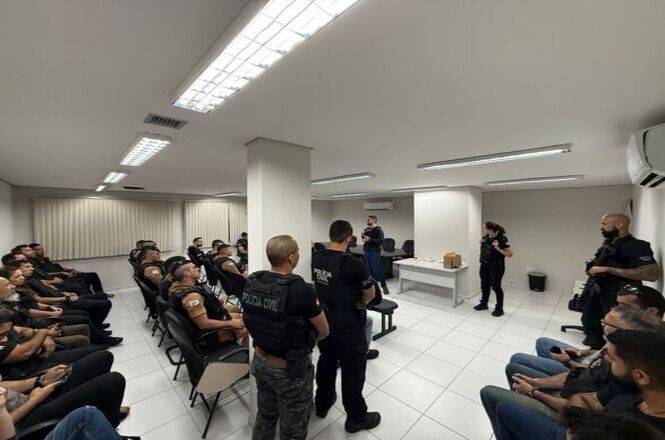 A imagem mostra uma sala de reuniões onde integrantes da Polícia Civil estão sentados em fileiras lado a lado. Todos estão uniformizados. Um homem e uma mulher estão em pé na frente da sala, de frente para todos. A sala é bem iluminada, com luzes fluorescentes no teto, e tem paredes brancas e piso de azulejo claro.