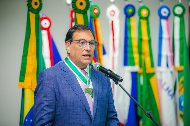 Foto horizontal: desembargador Francisco Oliveira Neto discursa ao microfone, com medalha no peito e bandeiras ao fundo, tons verde e amarelo.