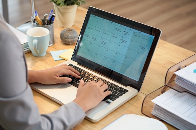 A imagem mostra uma pessoa usando um laptop em uma mesa de madeira clara. Na tela do laptop, há um calendário digital aberto. A pessoa está digitando no teclado. Ao lado, há uma caneca azul clara e alguns objetos de escritório. No canto direito da imagem, há uma pilha de papéis organizados em uma bandeja de documentos.