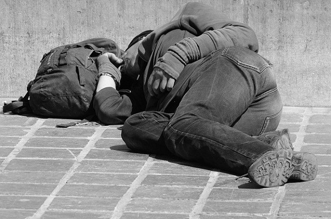 A imagem mostra uma pessoa deitada de lado em uma calçada, aparentemente dormindo ou descansando. A pessoa está vestindo calça jeans e um casaco com capuz. Ao lado dela, há uma mochila. A cena é em preto e branco, e a calçada é feita de blocos retangulares. A pessoa está deitada próxima a uma parede.
