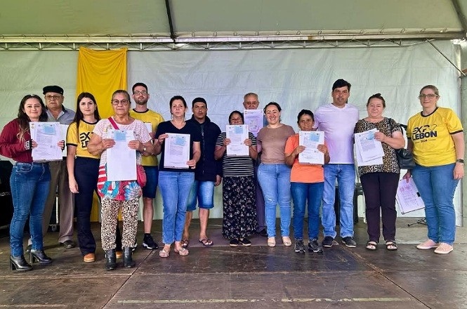 A imagem mostra um grupo de 13 pessoas em pé, posando para uma foto em um palco coberto. A maioria das pessoas está segurando documentos em suas mãos. O fundo é composto por uma cortina branca com um pedaço de tecido amarelo pendurado. Algumas pessoas estão vestidas com camisetas amarelas que têm o logotipo "AXX LEBON". O grupo parece estar participando de um evento ou cerimônia de entrega de documentos. As pessoas estão sorrindo e parecem felizes.