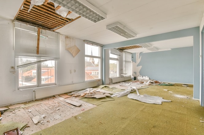 A imagem mostra uma sala grande e vazia em estado de abandono. O teto está danificado, com partes caindo e expostas, revelando a estrutura de madeira por trás. Há luminárias retangulares embutidas no teto. O chão está coberto por um carpete verde desgastado e há destroços espalhados por toda a sala, incluindo pedaços de gesso e madeira.  As janelas são grandes, com persianas parcialmente quebradas, permitindo a entrada de luz natural.