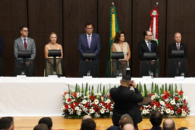 A imagem mostra uma cerimônia formal com seis pessoas em pé atrás de uma mesa coberta com uma toalha branca. Entre eles, o desembargador Francisco Oliveira Neto. Na frente da mesa, há um arranjo de flores vermelhas e brancas com folhas verdes. Todos estão vestidos formalmente. Ao fundo, há duas bandeiras, uma do Brasil e outra de Santa Catarina.