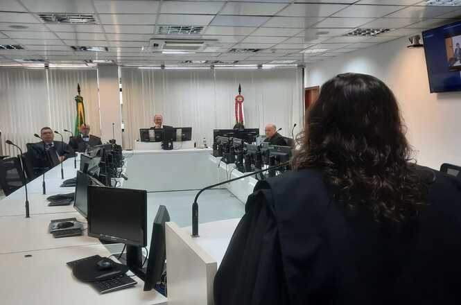 A imagem mostra uma sala de tribunal com várias pessoas presentes. No centro da imagem, há uma mesa em formato de "U" com computadores e microfones. Três homens estão sentados atrás dessa mesa, cada um em frente a um computador. Eles parecem ser juízes ou autoridades judiciais. À esquerda, há uma bandeira do Brasil e outra bandeira que não é totalmente visível. No primeiro plano, à direita, há uma pessoa de costas, com cabelos longos e vestindo uma toga preta, que parece estar se dirigindo aos juízes. No canto superior direito da imagem, há uma tela de televisão na parede. A sala tem um teto com iluminação embutida e janelas cobertas por persianas.