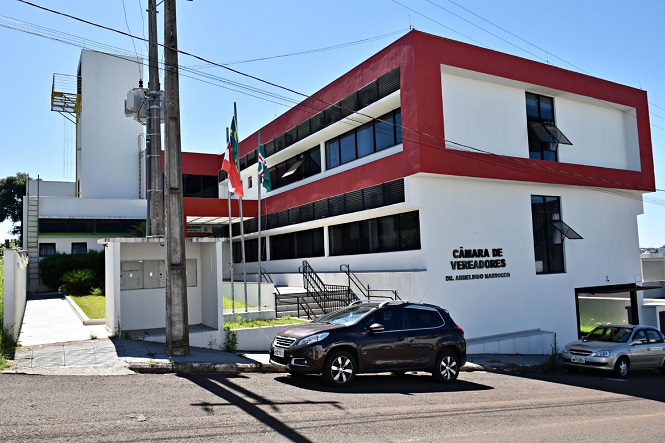 A imagem mostra um prédio moderno de dois andares com detalhes em vermelho e branco. Na fachada do prédio, está escrito "CÂMARA DE VEREADORES DR. AURÉLIO RIO MARQUES". Em frente ao prédio, há três mastros com bandeiras hasteadas. No estacionamento, há dois carros estacionados. O céu está claro e ensolarado, e há algumas árvores ao fundo.
