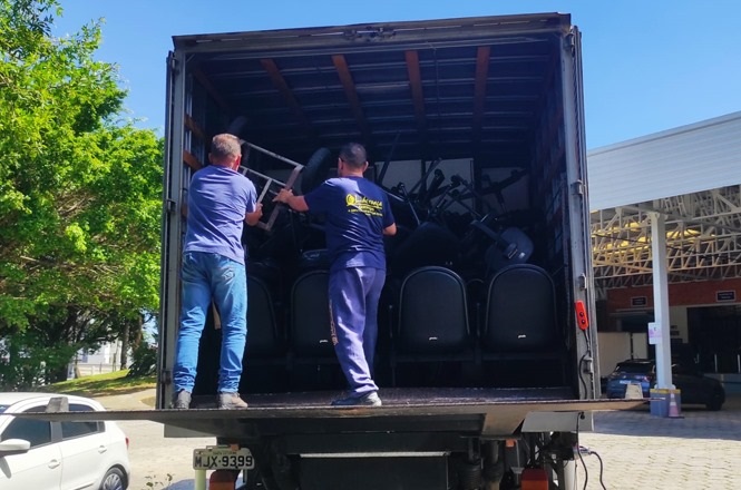 A imagem mostra a parte traseira de um caminhão aberto, onde dois homens estão carregando ou descarregando cadeiras. O interior do caminhão está cheio de cadeiras empilhadas. Os homens estão de costas para a câmera e usam camisetas azuis. O caminhão está estacionado em uma área externa, com árvores ao fundo e um edifício à direita.