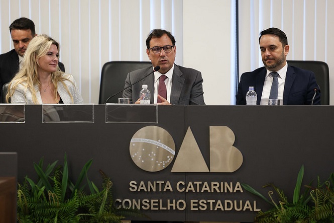 Foto horizontal: Francisco Oliveira Neto fala em evento da OAB Santa Catarina ao lado de autoridades em tons cinza.