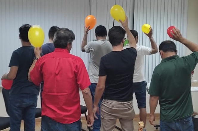 A imagem mostra um grupo de sete pessoas de costas. Cada pessoa está segurando um balão colorido acima da cabeça. Os balões são de cores variadas, incluindo amarelo, vermelho e laranja. As pessoas estão vestidas de forma casual, com camisetas e calças jeans. Ao fundo, há uma parede com persianas brancas fechadas.