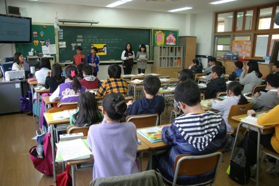A imagem mostra uma sala de aula com várias crianças sentadas em carteiras, voltadas para a frente da sala. Há um grupo de quatro pessoas em pé na frente, possivelmente professores ou alunos apresentando algo. A sala tem um quadro negro ao fundo, com algumas escritas e desenhos. As crianças parecem estar prestando atenção na apresentação. As carteiras estão organizadas em filas e há mochilas penduradas nas cadeiras. A sala é bem iluminada, com janelas à direita.