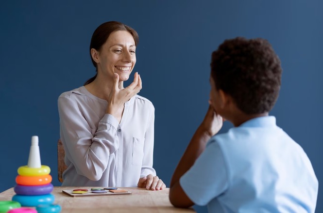 A imagem mostra uma mulher e um menino sentados frente a frente em uma mesa. A mulher, que tem cabelo castanho preso e veste uma blusa clara, está sorrindo e tocando seu rosto com a mão, como se estivesse demonstrando algo. O menino, que tem cabelo curto e encaracolado, está de costas para a câmera e parece estar prestando atenção na mulher. Na mesa, há alguns brinquedos coloridos, incluindo um conjunto de anéis empilháveis. O fundo é de cor azul sólida.