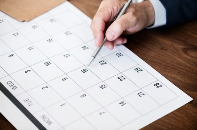 A imagem mostra uma mão segurando uma caneta sobre um calendário de mesa. O calendário está aberto no mês de abril, com os dias organizados em uma grade. A mão parece estar prestes a escrever ou marcar algo no dia 16. O calendário está sobre uma superfície de madeira.