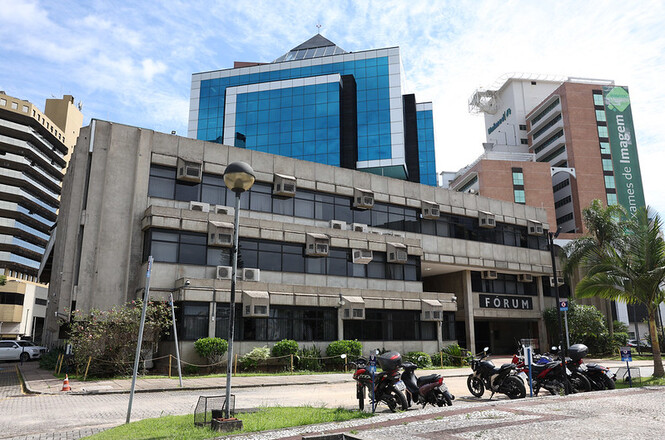 A imagem mostra um edifício de aparência oficial, identificado como "FÓRUM" na entrada. O prédio é de concreto, com várias janelas e unidades de ar-condicionado visíveis. Na frente do edifício, há algumas motos estacionadas em um espaço pavimentado. Ao fundo, há outros prédios mais altos, um deles com fachada de vidro azul e outro com uma faixa verde vertical com o texto "Exames de Imagem". O céu está claro, com algumas nuvens. Há também uma área com grama e algumas plantas próximas ao fórum.
