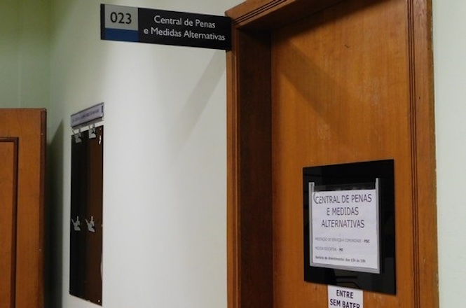A imagem mostra um corredor de um prédio com uma porta de madeira à direita. Acima da porta, há uma placa com o número "023" e o texto "Central de Penas e Medidas Alternativas". Na porta, há um aviso que diz "CENTRAL DE PENAS E MEDIDAS ALTERNATIVAS" e abaixo, em letras menores, "PROTEÇÃO SOCIAL E CIDADANIA - PMC NÚCLEO REGIONAL". Mais abaixo, há um outro aviso que diz "ENTRE SEM BATER". Ao lado da porta, na parede, há um quadro de avisos com alguns papéis presos.