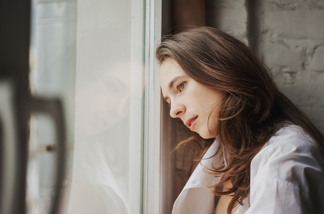 A imagem mostra uma mulher segurando um microfone e falando em um ambiente que parece ser uma sala de conferências ou reunião. Ela está vestindo um casaco cinza e um cachecol colorido. Atrás dela, há um banner com uma imagem de mãos e texto parcialmente visível que diz: "Sinal vermelho contra a violência doméstica". O banner também possui um logotipo e um QR code na parte inferior. Ao fundo, há cadeiras e mesas de escritório.