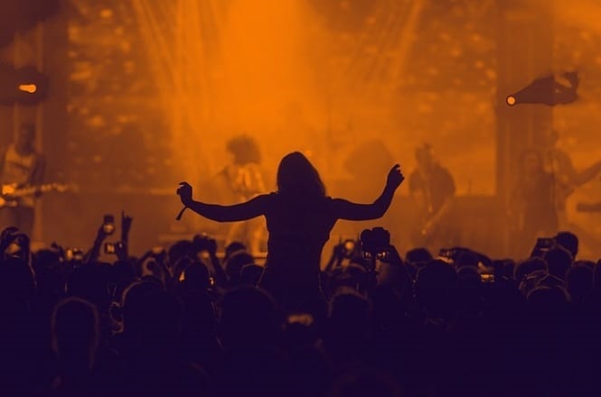 A imagem mostra uma cena de um show ou concerto. No primeiro plano, há uma multidão de pessoas, muitas delas segurando celulares para gravar ou tirar fotos do evento. No centro da imagem, destaca-se a silhueta de uma pessoa com os braços levantados, possivelmente dançando ou celebrando a música. Ao fundo, no palco, é possível ver músicos tocando instrumentos, mas eles estão desfocados. A iluminação é predominantemente em tons de laranja, criando uma atmosfera vibrante e energética.