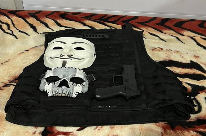 A imagem mostra um colete tático preto sobre uma superfície que parece ser uma coberta com estampa de tigre. Em cima do colete, há uma máscara branca do tipo "Guy Fawkes", popularizada pelo filme "V de Vingança", uma máscara de caveira prateada e uma pistola preta. A máscara de Guy Fawkes tem um bigode e um cavanhaque estilizados. A máscara de caveira tem um design que imita um crânio humano. A pistola está posicionada ao lado das máscaras.