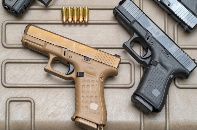 A imagem mostra duas pistolas e algumas balas. A pistola à esquerda é de cor marrom claro, enquanto a pistola à direita é preta. Ambas estão posicionadas sobre uma superfície com um padrão de linhas retangulares. Acima das pistolas, há um conjunto de seis balas dispostas em linha. As pistolas têm um design moderno e parecem ser de um modelo semelhante, mas com cores diferentes.