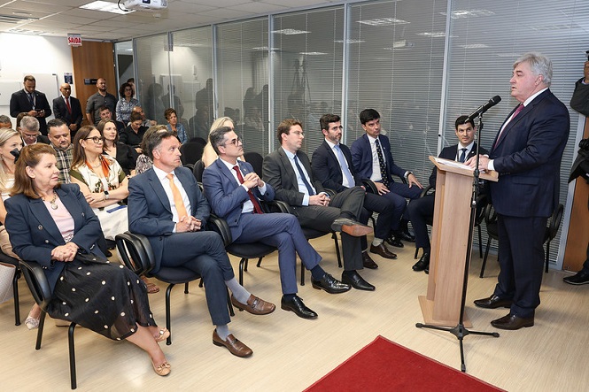 A imagem mostra uma sala de conferências onde várias pessoas estão sentadas, prestando atenção em um homem que está em pé, falando ao microfone em um púlpito. O homem que está falando veste um terno escuro e gravata vermelha. Entre o público, há homens e mulheres, a maioria vestindo trajes formais como ternos e blazers. A sala tem paredes de vidro e há uma placa de "SAÍDA" visível ao fundo. As pessoas parecem estar em um evento formal ou reunião.