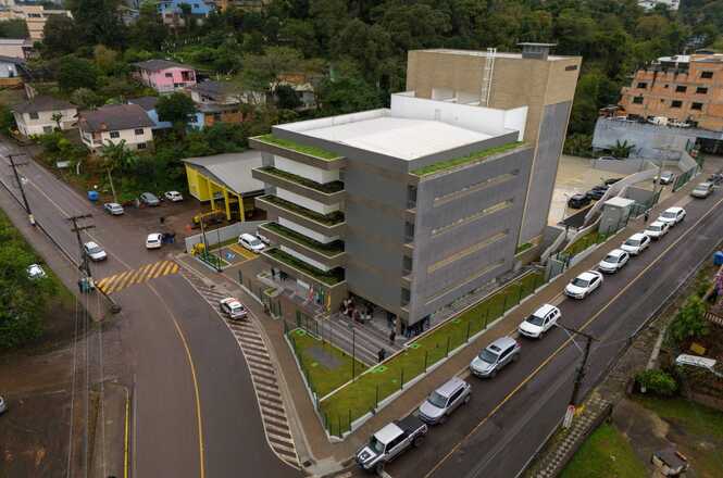 A imagem mostra uma vista aérea de um edifício moderno de vários andares, localizado em uma esquina. O prédio tem um design contemporâneo com linhas retas e varandas em cada andar, algumas delas com vegetação. O telhado também possui áreas verdes. Ao redor do edifício, há uma calçada e uma cerca baixa. Na rua ao lado do prédio, há uma fila de carros estacionados. A área ao redor parece ser uma zona urbana com algumas casas e edifícios comerciais. Há também uma estrada que faz uma curva acentuada ao lado do prédio. A vegetação e algumas árvores são visíveis ao fundo, sugerindo que a área tem um pouco de verde.