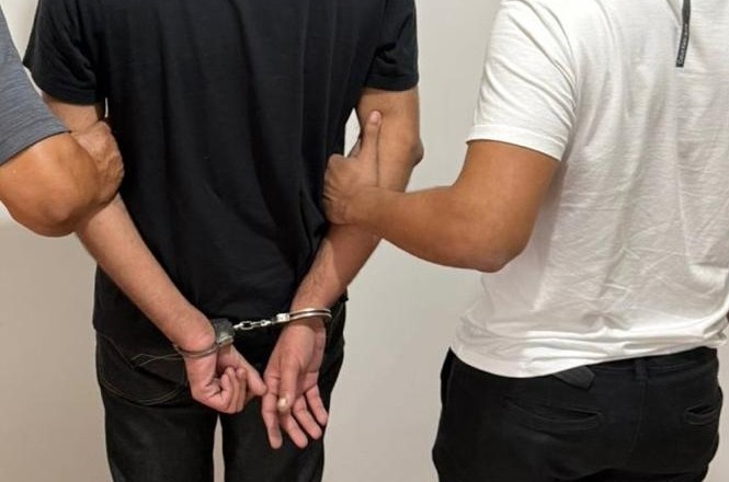 A imagem mostra três homens de costas. O homem no centro está algemado com as mãos para trás. Ele veste uma camiseta preta e calça escura. Os outros dois homens, um à esquerda e outro à direita, estão segurando os braços do homem algemado. O homem à esquerda veste uma camiseta cinza, e o homem à direita veste uma camiseta branca. A cena parece ocorrer em um ambiente interno com uma parede clara ao fundo.