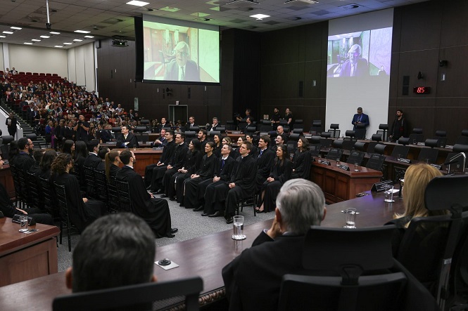 A imagem mostra uma cerimônia formal em um auditório. No centro, há várias pessoas sentadas em cadeiras, todas vestindo togas pretas, sugerindo que são juízes ou formandos em uma cerimônia de graduação. À frente deles, há uma mesa comprida com mais pessoas sentadas, também em trajes formais. No fundo, há um grande público sentado em cadeiras vermelhas. Duas grandes telas de projeção mostram a imagem de um homem falando, provavelmente um discurso ou apresentação. O ambiente é bem iluminado e parece ser uma sala de tribunal ou um auditório de conferências.