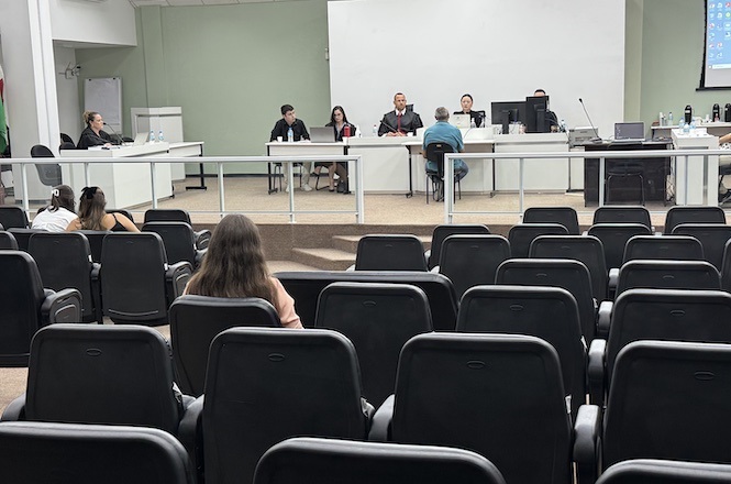 A imagem mostra uma sala de audiências ou tribunal. Na parte frontal, há uma mesa comprida onde estão sentadas cinco pessoas, possivelmente advogados, juízes ou participantes de uma audiência. Atrás deles, há uma parede com um grande quadro branco ou tela de projeção. À direita, há uma tela de computador projetada na parede. No primeiro plano, há várias fileiras de cadeiras pretas, algumas das quais estão ocupadas por pessoas assistindo à sessão. A sala tem um ambiente formal e organizado, típico de um tribunal ou sala de conferências.