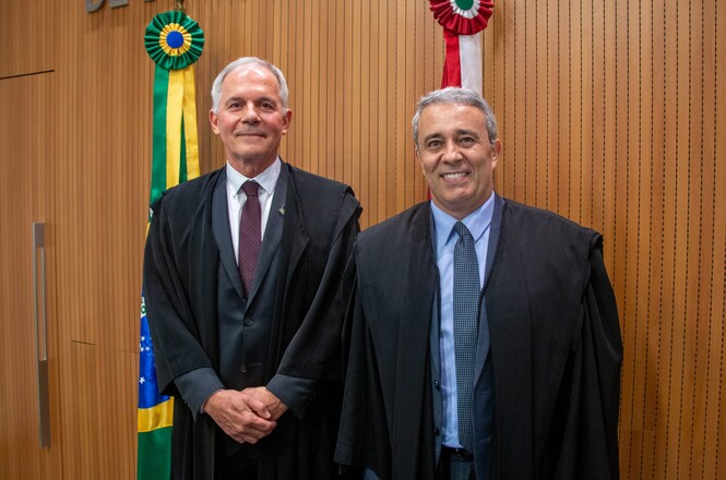 A imagem mostra dois homens de pé, lado a lado, ambos vestindo togas pretas. O homem à esquerda tem cabelo grisalho, está usando uma camisa branca e gravata vinho. O homem à direita também tem cabelo grisalho, está usando uma camisa azul clara e gravata azul escura. Atrás deles, há duas bandeiras: a bandeira do Brasil à esquerda e uma bandeira com listras verticais vermelhas e brancas à direita. O fundo é composto por uma parede de madeira com ripas verticais. Ambos os homens estão sorrindo.