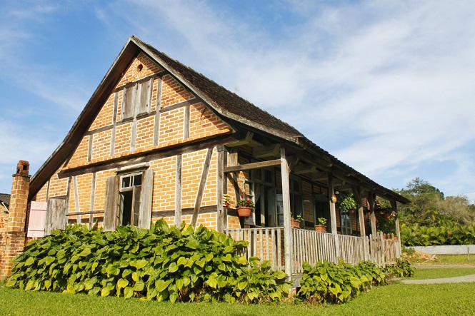 A imagem mostra uma casa de estilo enxaimel, típica da arquitetura germânica, construída com tijolos e madeira. A casa tem um telhado inclinado coberto por telhas escuras. Na frente, há um jardim com plantas verdes e uma varanda com vasos de flores pendurados. O céu está claro, com poucas nuvens, e o ambiente parece ser de uma área rural ou de campo, com bastante vegetação ao redor.