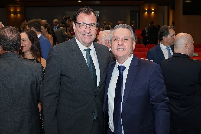 A imagem mostra duas pessoas em destaque, ambas usando ternos. O homem à esquerda é mais alto, usa óculos e está vestindo um terno cinza escuro com uma gravata azul. Ele está sorrindo. O homem à direita também está sorrindo e veste um terno azul escuro com uma gravata azul. Eles estão em um ambiente fechado, que parece ser um auditório ou sala de conferências, com várias outras pessoas ao fundo, algumas sentadas em cadeiras vermelhas. O ambiente está bem iluminado e há uma atmosfera de evento formal ou reunião.