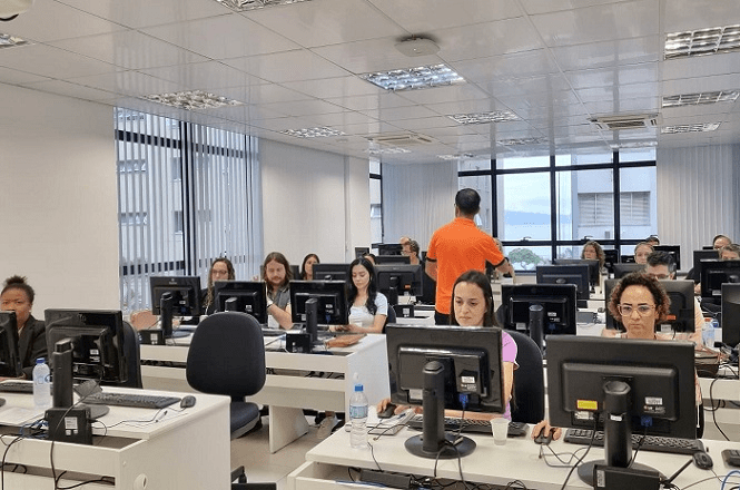 A imagem mostra uma sala de aula de informática com várias pessoas sentadas em frente a computadores. A sala é bem iluminada, com janelas grandes ao fundo. As pessoas estão concentradas nas telas dos computadores. Há um homem de pé, vestindo uma camisa laranja, que parece estar instruindo ou ajudando os alunos. As mesas são brancas e cada uma tem um monitor, teclado e mouse. Há também garrafas de água sobre as mesas. A sala tem um teto com iluminação embutida e o ambiente parece ser moderno e organizado.