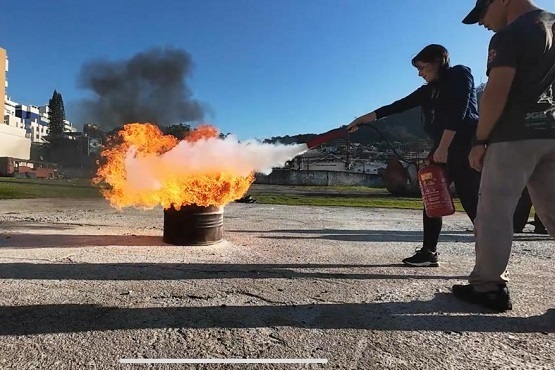 A imagem mostra uma pessoa usando um extintor de incêndio para apagar um fogo que está saindo de um tambor de metal. O fogo está produzindo chamas laranja e fumaça preta. A pessoa está vestida com roupas escuras e está segurando o extintor com as duas mãos, direcionando o jato de espuma ou pó químico para as chamas. Ao lado, há outra pessoa observando a ação. O cenário parece ser ao ar livre, em um espaço aberto com algumas construções ao fundo e um céu azul claro.