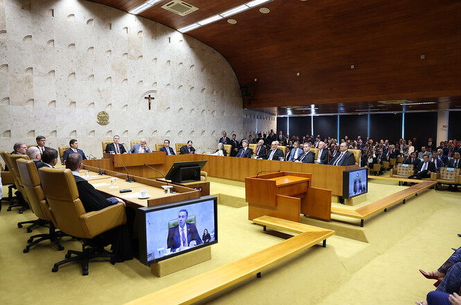 A imagem mostra uma sessão no plenário do Supremo Tribunal Federal (STF) do Brasil. O ambiente é amplo, com um teto de madeira e paredes claras. Há várias pessoas sentadas em cadeiras dispostas em formato de "U", com um grupo de ministros do STF em cadeiras maiores e mais próximas ao centro. Na frente deles, há mesas com microfones e monitores. No centro da imagem, há um crucifixo na parede. O público está sentado em cadeiras ao fundo, assistindo à sessão. Há também duas telas grandes que exibem a imagem de uma pessoa falando, possivelmente uma transmissão ao vivo ou uma video conferência.
