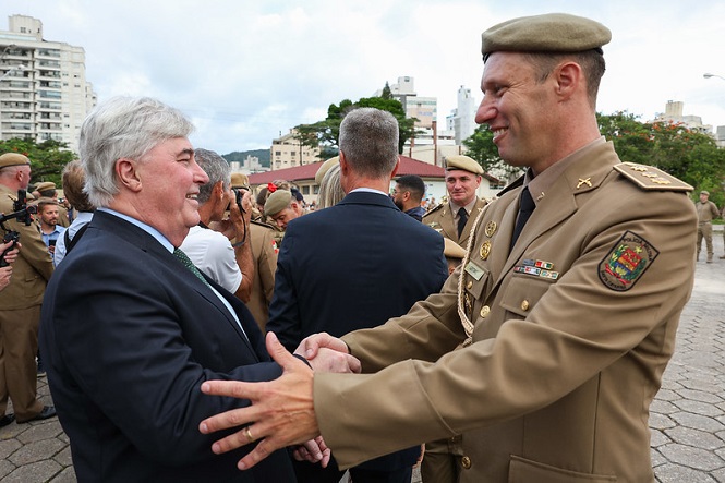 A imagem mostra duas pessoas se cumprimentando com um aperto de mão. À esquerda, há um homem de cabelos grisalhos, vestindo um terno escuro com uma camisa branca e gravata verde. À direita, há um homem usando um uniforme militar bege, com várias insígnias e um distintivo no braço. Ele também está usando um quepe combinando com o uniforme. Ao fundo, há várias outras pessoas, algumas também em uniformes militares, em um ambiente ao ar livre com prédios e árvores ao fundo. Parece ser um evento formal ou cerimônia militar.