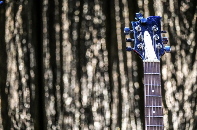 A imagem mostra o braço de uma guitarra elétrica, focando na parte superior onde estão as tarraxas. A guitarra tem um acabamento brilhante e escuro, possivelmente azul, e as tarraxas são metálicas. O fundo da imagem é desfocado, com um padrão de luzes que parecem ser reflexos, criando um efeito visual interessante. A imagem destaca o contraste entre a nitidez da guitarra e o fundo desfocado