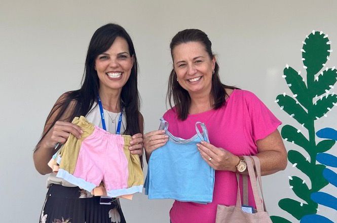 A imagem mostra duas mulheres sorrindo e segurando roupas infantis. A mulher à esquerda tem cabelo escuro e veste uma blusa clara com um crachá pendurado no pescoço. Ela segura um par de shorts infantis, um rosa e outro amarelo. A mulher à direita tem cabelo castanho claro e veste uma blusa rosa. Ela segura uma camiseta infantil azul. Ao fundo, há uma parede clara com uma decoração que parece ser uma planta estilizada em verde e azul. Ambas parecem estar em um ambiente iluminado e estão felizes.