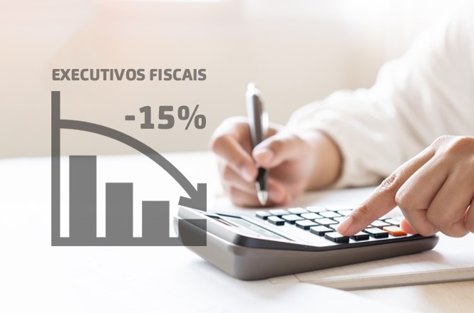 A imagem mostra uma pessoa usando uma calculadora, com uma caneta na mão, enquanto faz anotações em um papel. Sobreposta à imagem, há um gráfico de barras com uma seta descendente, indicando uma queda. Acima do gráfico, está escrito "EXECUTIVOS FISCAIS" e, ao lado, "-15%". Isso sugere uma redução de 15% em algum aspecto relacionado a executivos fiscais.