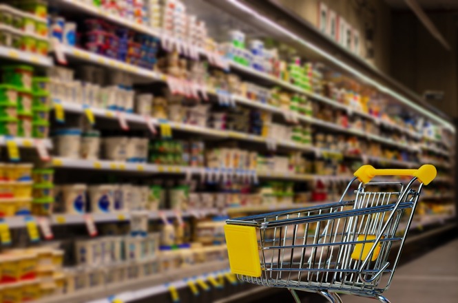 A imagem mostra o interior de um supermercado. Em primeiro plano, há um carrinho de compras vazio com detalhes amarelos na alça e na lateral. Ao fundo, há prateleiras cheias de produtos diversos, como alimentos e produtos embalados. As prateleiras estão bem organizadas e há etiquetas de preços visíveis, mas os detalhes dos produtos e preços não são legíveis devido ao desfoque da imagem.