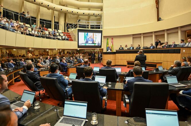 A imagem mostra uma sessão em um plenário legislativo. Há várias pessoas sentadas em cadeiras ao redor de mesas em formato semicircular, todas voltadas para um púlpito central onde uma pessoa está de pé, aparentemente discursando. Na parte superior da imagem, há um telão exibindo uma pessoa falando, possivelmente transmitindo uma mensagem ou discurso. O ambiente é bem iluminado e há bandeiras ao lado do púlpito, indicando que se trata de um evento formal. As pessoas presentes parecem estar prestando atenção ao que está sendo dito. Além disso, há um público sentado em uma galeria superior, observando a sessão.