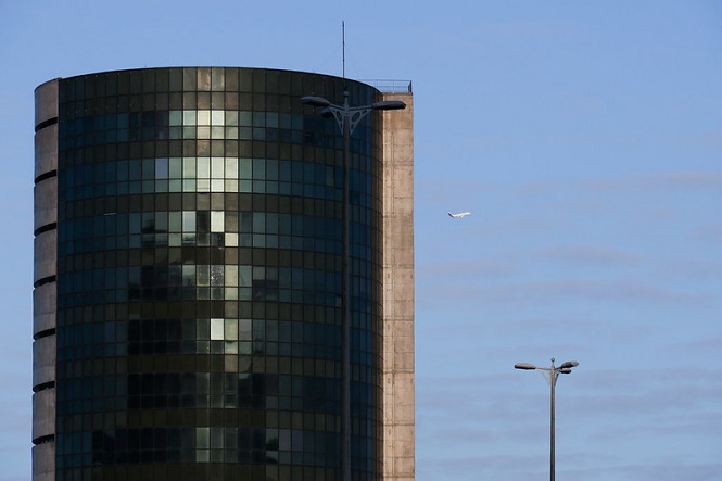 A imagem mostra um edifício alto com fachada de vidro, refletindo a luz do sol. O prédio tem uma forma cilíndrica e está localizado à esquerda da imagem. À direita, no céu azul, há um avião pequeno voando. Na parte inferior direita da imagem, há dois postes de iluminação. O céu está claro, com algumas nuvens finas.