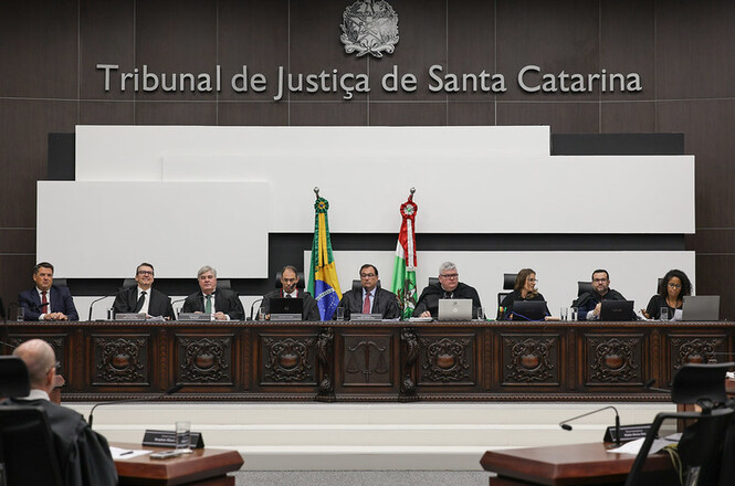 A imagem mostra uma sessão no Tribunal de Justiça de Santa Catarina. Há uma mesa comprida com nove pessoas sentadas, todas de terno ou vestes formais, possivelmente juízes ou desembargadores. Atrás deles, na parede, está escrito "Tribunal de Justiça de Santa Catarina" e há um brasão acima do texto. Na frente da mesa, há duas bandeiras: a do Brasil e a de Santa Catarina. A sala parece ser um auditório ou plenário, com outras pessoas sentadas em mesas menores em primeiro plano.